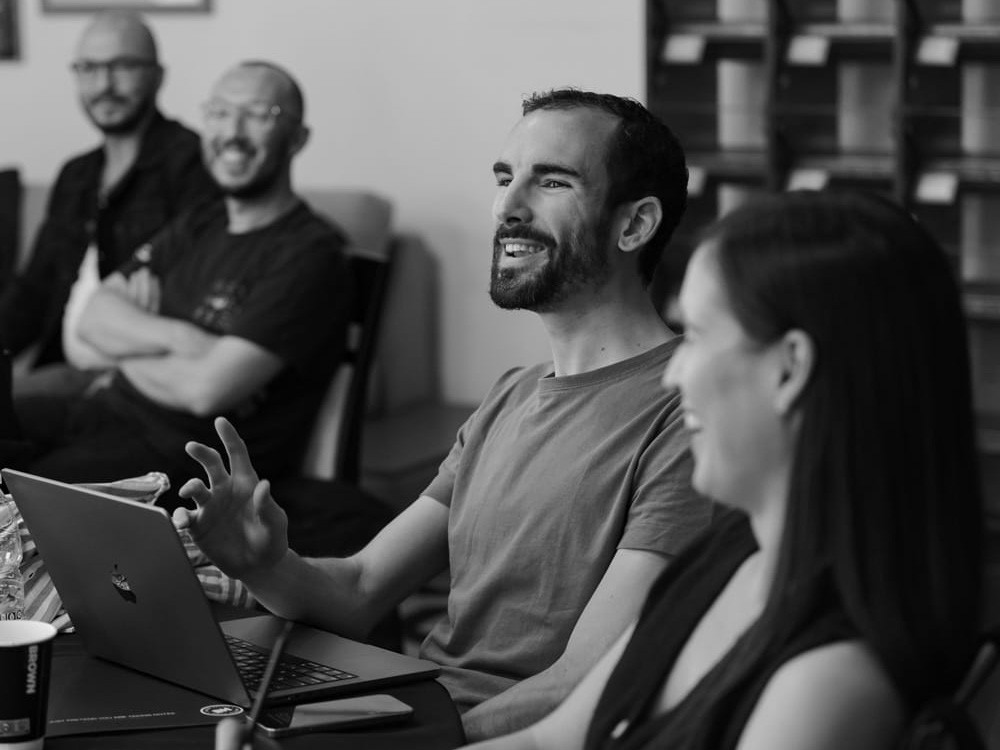 A photograph of three GitBook team members sitting around a table listening to a presentation. One team member has his laptop open